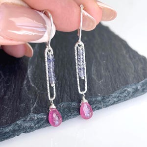 May include: Silver drop earrings featuring a vertical design with a row of small, light purple beads. The earrings have a teardrop-shaped, pink gemstone at the bottom. The earrings are displayed against a dark, textured background.