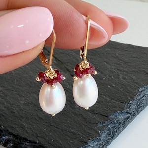 May include: Gold-toned drop earrings featuring a cluster of small red gemstones above a pear-shaped white pearl. The earrings are held up against a dark, textured surface.