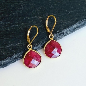 May include: A pair of gold-colored leverback earrings with faceted, teardrop-shaped, deep red gemstones. The earrings are displayed on a dark, textured surface with a white background.