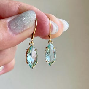May include: A pair of gold dangle earrings with light blue marquise-shaped gemstones. The earrings have a simple design and are perfect for everyday wear.