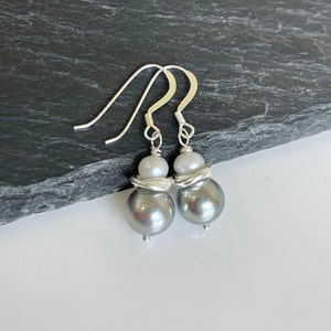 May include: A pair of silver drop earrings featuring gray and white pearls. The earrings have a large gray pearl at the bottom, a smaller white pearl, and a silver wire design. The hooks are silver.