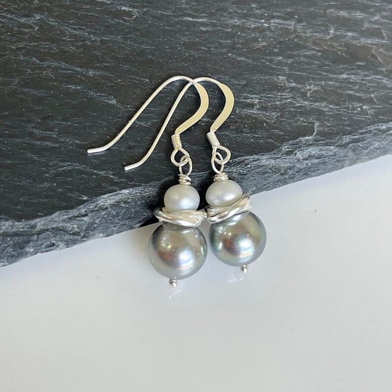 May include: A pair of silver drop earrings featuring gray and white pearls. The earrings have a large gray pearl at the bottom, a smaller white pearl, and a silver wire design. The hooks are silver.