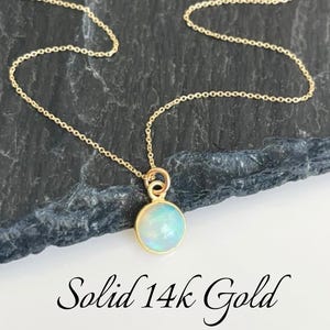 May include: A delicate gold chain necklace with a small, round, light blue opal pendant. The necklace is made of solid 14k gold.