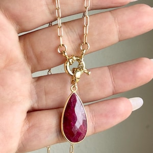 May include: A gold-toned necklace featuring a faceted, teardrop-shaped deep red gemstone pendant. The pendant is attached to a gold-toned chain by a circular toggle clasp mechanism. The chain consists of elongated, rectangular-shaped links.