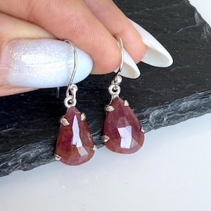 May include: A pair of drop earrings featuring faceted, pear-shaped dark pink gemstones set in silver-toned metal prongs. Each earring hangs from a simple silver-toned hook wire, showcasing the translucent, multi-faceted surface of the stones.