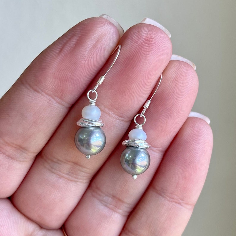 May include: A pair of dangle earrings featuring a dark gray pearl, a light gray bead, and silver-tone metal accents. The earrings have a simple, elegant design with a hook closure. The earrings are shown against a neutral background.