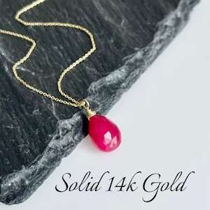 May include: A delicate gold chain necklace with a bright pink gemstone pendant. The pendant is faceted and has a teardrop shape. The text "Solid 14k Gold" is visible in the image.