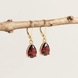 May include: A pair of gold leverback earrings featuring faceted, pear-shaped deep red garnet gemstones. The earrings are suspended from a thin, horizontal wooden branch, showcasing the polished metal settings and the translucent quality of the stones.