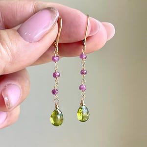 May include: Gold-tone dangle earrings featuring a series of small, round, pink gemstones and a teardrop-shaped, light green gemstone at the bottom. The earrings are suspended from a gold-colored hook.