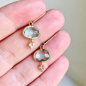 K&ouml;nnte beinhalten: Goldene Ohrh&auml;nger mit einem hellblauen ovalen Edelstein und einem Cluster aus drei kleinen klaren Edelsteinen.
