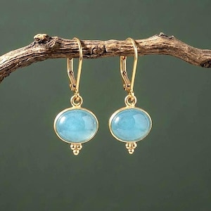 May include: A pair of gold-toned drop earrings featuring oval-shaped, light blue cabochon stones. Each earring is suspended from a gold-toned leverback closure and includes a small, decorative three-bead detail at the base of the stone setting.