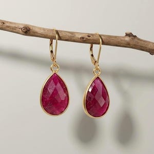 May include: A pair of gold leverback earrings featuring faceted, teardrop-shaped deep pink gemstones. The earrings are suspended from a thin, horizontal wooden branch, showcasing the vibrant color and geometric cut of the stones clearly.