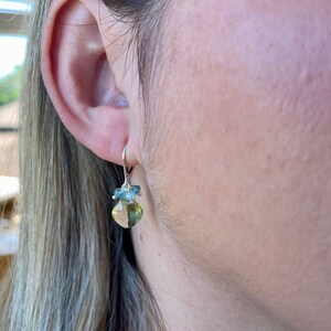 May include: A pair of silver dangle earrings with a light green and blue gemstone pendant. The pendant is a square shape with rounded edges and is surrounded by smaller blue gemstones.