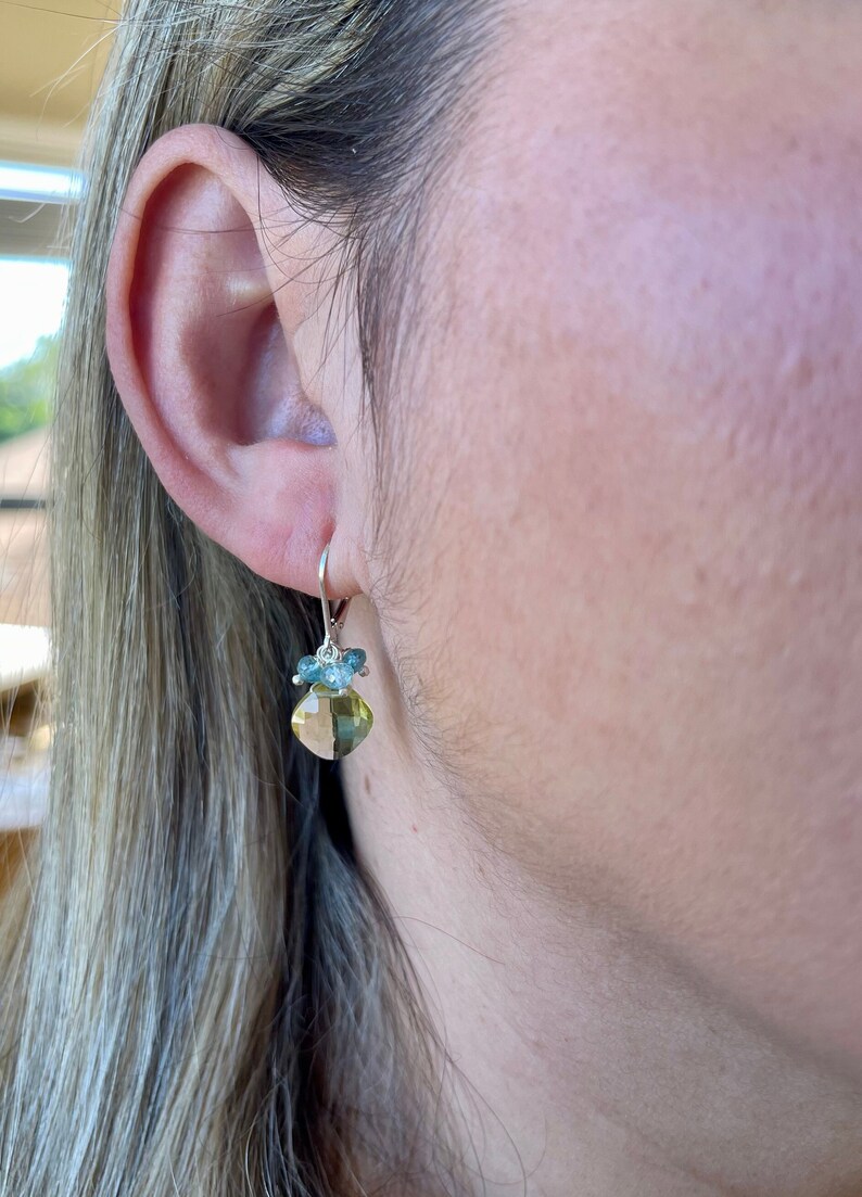May include: A pair of silver dangle earrings with a light green and blue gemstone pendant. The pendant is a square shape with rounded edges and is surrounded by smaller blue gemstones.