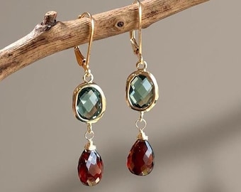 Pode incluir: Um par de brincos de ouro com fecho de alavanca, apresentando uma pedra preciosa verde oval facetada no topo e uma pedra vermelha escura facetada em forma de lágrima pendurada abaixo. Os brincos estão suspensos num ramo de madeira fino.