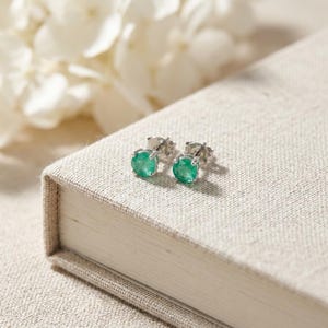 May include: A pair of round stud earrings featuring vibrant green gemstones set in polished silver-toned metal prongs. The earrings are positioned on a textured, light-colored surface, showcasing a classic design with a secure post backing.