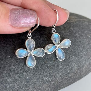May include: A pair of silver-toned earrings featuring a four-petal flower design. Each petal is a teardrop shape, filled with a translucent, iridescent blue-white stone. The earrings are held up by a silver hook.