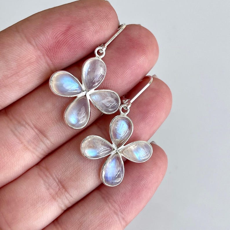 May include: A pair of silver earrings featuring four-petal flower designs. Each petal is a clear, iridescent stone with blue and white hues. The earrings are held by a person's hand.