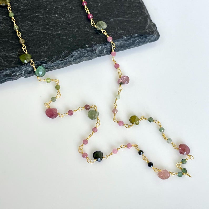Pode incluir: Um colar longo de corrente dourada com gemas rosa, verdes e pretas. O colar &eacute; feito de pequenas contas douradas e gemas maiores de forma irregular.