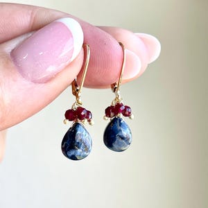 May include: A pair of gold-colored leverback earrings. Each earring features a cluster of small, red beads above a teardrop-shaped, dark blue stone. The earrings are held up by a person's fingers.