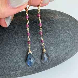 May include: Gold-toned dangle earrings featuring a series of small, round, red beads and faceted, diamond-shaped blue-gray stones. The earrings are suspended from gold-colored ear wires.