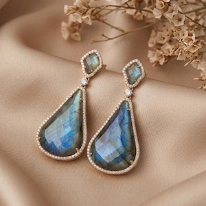 May include: A pair of drop earrings featuring faceted, pear-shaped labradorite stones with blue iridescence. Each earring has a smaller, diamond-shaped labradorite top piece, connected by a tiny clear stone, all set in a gold-toned metal frame.