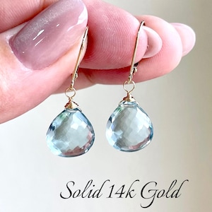 May include: A pair of gold-colored drop earrings with teardrop-shaped, faceted, light blue gemstones. The earrings are suspended from gold-colored hooks. The text "Solid 14k Gold" is written in cursive at the bottom of the image.