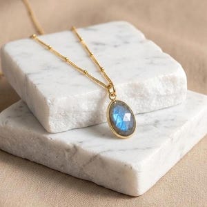 May include: A gold necklace featuring a delicate chain with small beads and an oval-shaped labradorite pendant. The gemstone displays a faceted surface with iridescent blue flashes, securely set within a polished gold-toned metal frame.