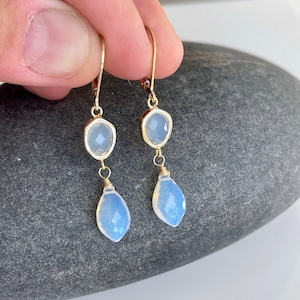 May include: A pair of gold dangle earrings with two teardrop-shaped opal stones. The top stone is a faceted square and the bottom stone is a teardrop.