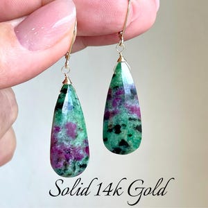 May include: A pair of 14k gold teardrop earrings with green and pink gemstone drops. The earrings are shown on a hand.