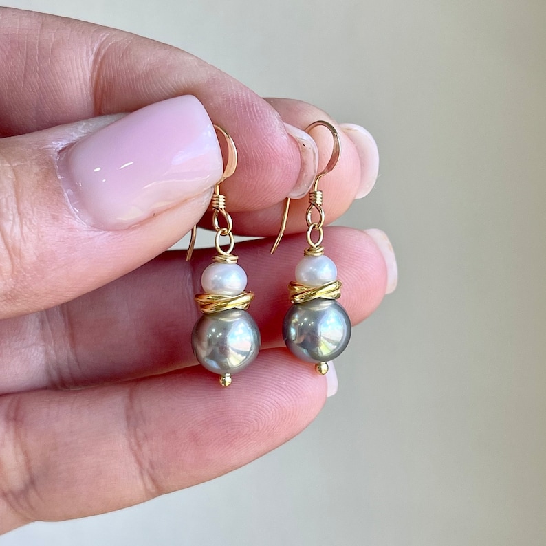 May include: A pair of gold-colored drop earrings featuring gray and white pearls. The earrings have a gold-colored hook and are composed of a large gray pearl, a gold-colored accent, and a smaller white pearl. The earrings are held in a hand.