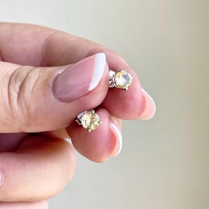 May include: A pair of stud earrings featuring round, light yellow gemstones set in silver-colored metal. The earrings are small and delicate, with a simple, classic design. The gemstones have a bright, clear appearance.