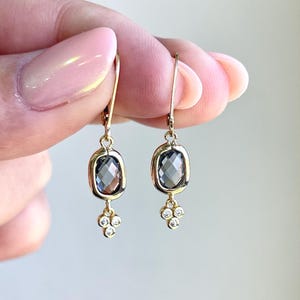 May include: A pair of gold-colored drop earrings. Each earring features a dark gray, oval-shaped stone set in a gold frame. Below the stone, three small, round, clear stones are clustered together.