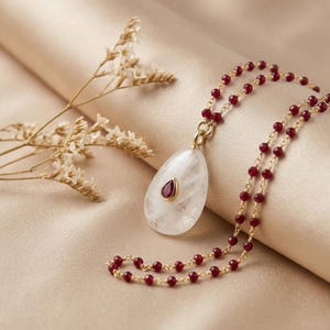 May include: A necklace featuring a teardrop-shaped white stone pendant with a small, central, teardrop-shaped red gemstone inlay. The chain is composed of small, faceted, deep red beads linked together with thin gold-toned metal wire.