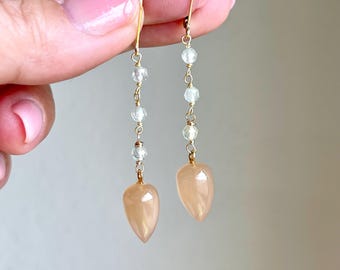 May include: A pair of long, delicate earrings with a teardrop-shaped peach-colored gemstone at the bottom. The earrings are made with a gold chain and small, clear gemstones.