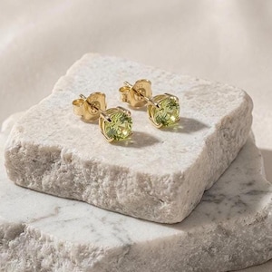 May include: A pair of gold stud earrings featuring round-cut, light green gemstones held in place by four-prong settings. The earrings are designed with a post-back mechanism, showcasing a bright, reflective metallic finish and clear, faceted stones.