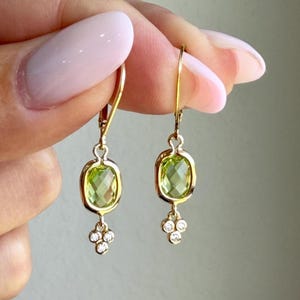 May include: A pair of gold drop earrings featuring a faceted, oval-shaped light green gemstone set in a gold frame. Below each oval stone is a small cluster of three tiny, round, clear white stones arranged in a triangular pattern.