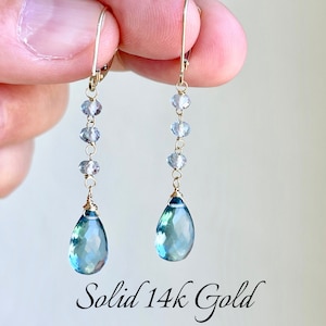 May include: A pair of dangle earrings with a gold-colored metal. Each earring features a series of small, light blue beads leading to a larger, teardrop-shaped blue gemstone. The text "Solid 14k Gold" is visible.