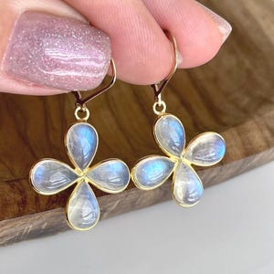 May include: Gold-tone earrings featuring four-petal flower designs. Each petal is a teardrop shape with a light blue, iridescent stone. The earrings are held up by a person's hand.