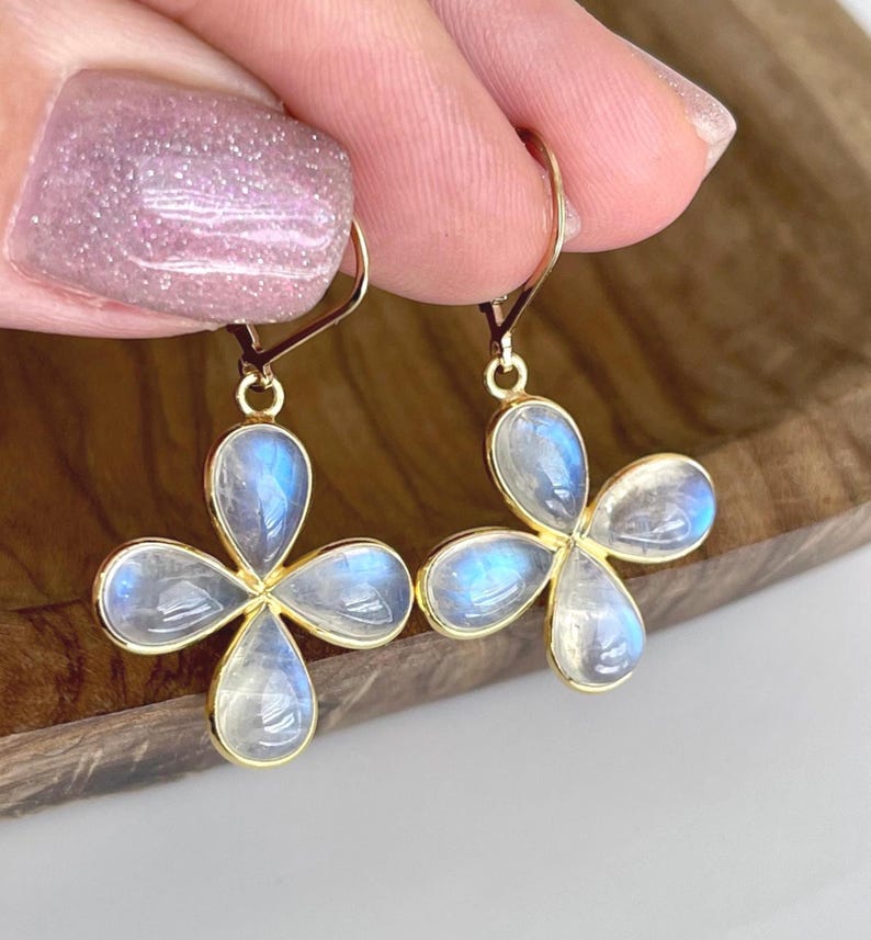 May include: Gold-tone earrings featuring four-petal flower designs. Each petal is a teardrop shape with a light blue, iridescent stone. The earrings are held up by a person's hand.
