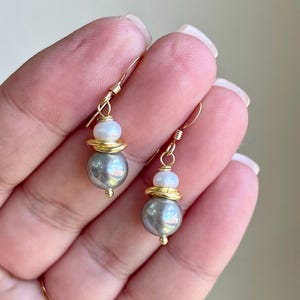 May include: A pair of gold-colored drop earrings. Each earring features a gray pearl, a gold-colored ring, and a white pearl. The earrings are suspended from gold-colored hooks.