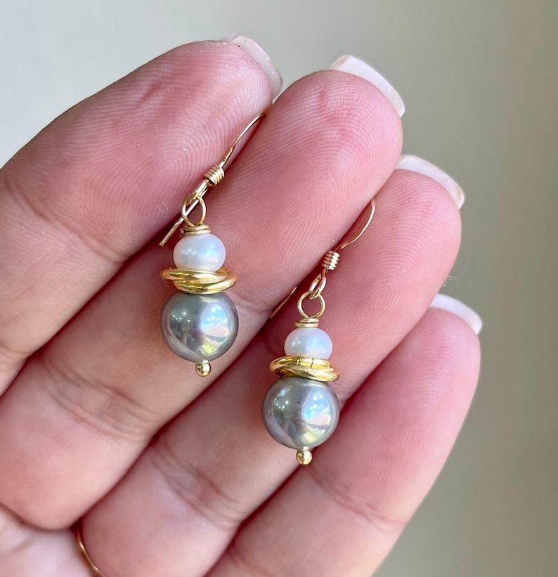 May include: A pair of gold-colored drop earrings. Each earring features a gray pearl, a gold-colored ring, and a white pearl. The earrings are suspended from gold-colored hooks.