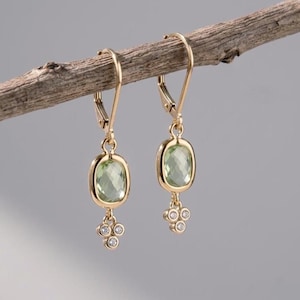 May include: A pair of gold drop earrings featuring a faceted light green cushion-cut gemstone in a bezel setting, suspended from a lever-back closure, with a small triangular cluster of three tiny clear round stones dangling at the bottom.
