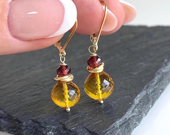Könnte beinhalten: Ein Paar Ohrhänger mit facettierten, goldgelben Citrinperlen und kleineren, dunkelroten Granatperlen. Die Steine hängen an goldfarbenen Brisur-Verschlüssen, wobei jede Perle mit einem kleinen goldfarbenen Metallring und Draht verziert ist.