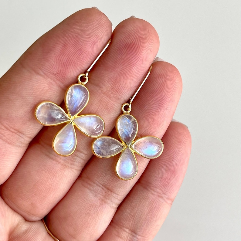 May include: A pair of gold-colored earrings featuring a four-petal flower design. Each petal is a translucent, iridescent stone with a gold border. The earrings are held up by a gold hook.