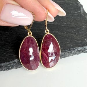May include: A pair of drop earrings featuring faceted, teardrop-shaped deep red ruby gemstones set in gold-toned metal bezels. The earrings are attached to gold-toned lever-back closures, showcasing a polished finish and a distinct geometric cut.