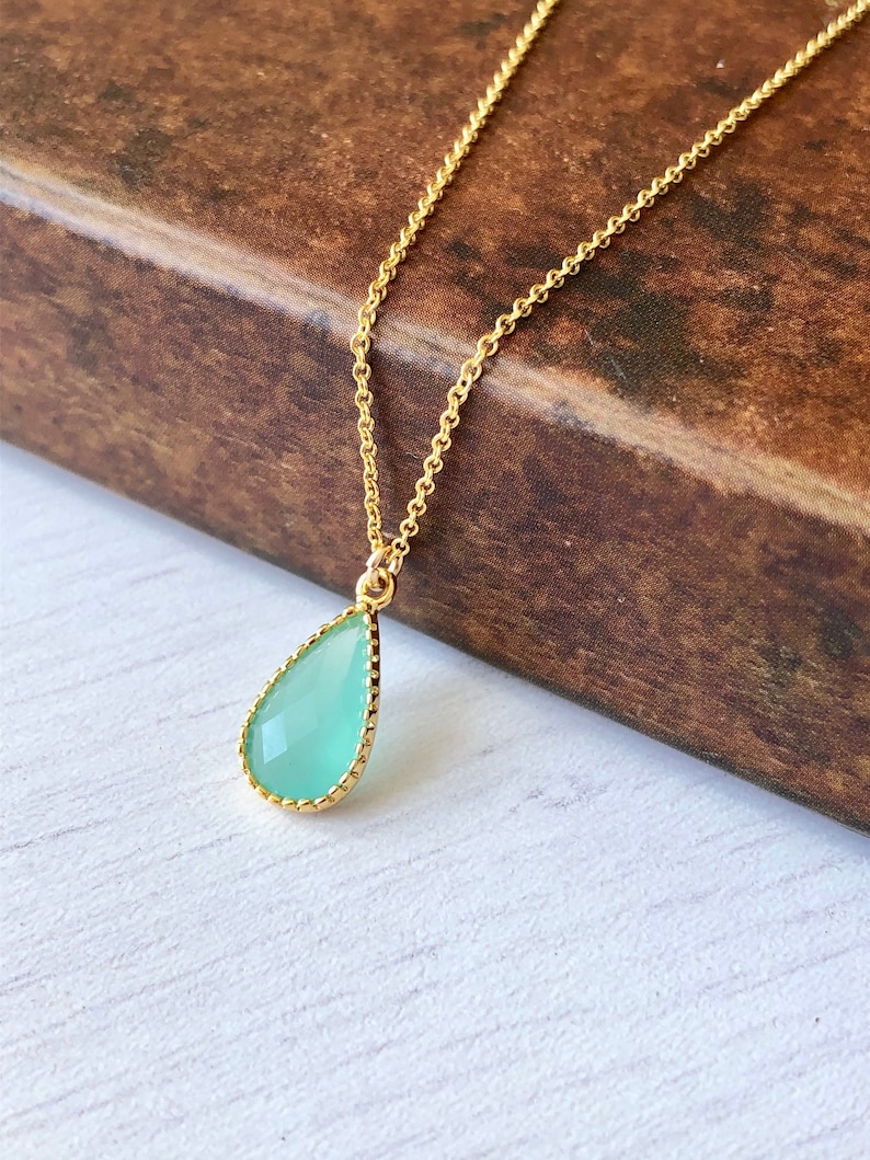 Puede incluir: Un delicado collar de cadena dorada con un colgante de piedra preciosa verde en forma de l&aacute;grima. El colgante tiene un borde dorado.