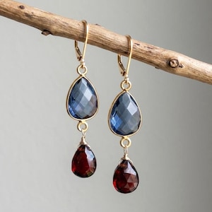 May include: A pair of gold-tone dangle earrings featuring a faceted, pear-shaped blue stone suspended above a smaller, faceted, pear-shaped deep red stone. The earrings are attached to gold-tone lever-back closures, showcasing a layered design.