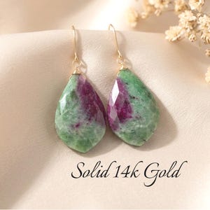 May include: A pair of teardrop-shaped ruby zoisite earrings featuring faceted green and magenta stones suspended from thin gold hooks. The text Solid 14k Gold is printed below the earrings, indicating the material of the metal components.