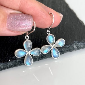 May include: A pair of silver-toned earrings featuring a four-petal flower design. Each petal holds a pear-shaped, iridescent, blue-white stone. The earrings are held up by a person's fingers against a dark background.